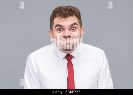 funny portrait of young man in white shirt and tie standing blowing his cheeks and looking at camera with funny face and big eyes. indoor isolated on Stock Photo