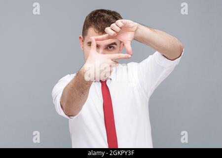 Portrait of young handsome attentive man in white shirt and tie standing looking through photo frame made of hands, focusing and cropping image. indoo Stock Photo