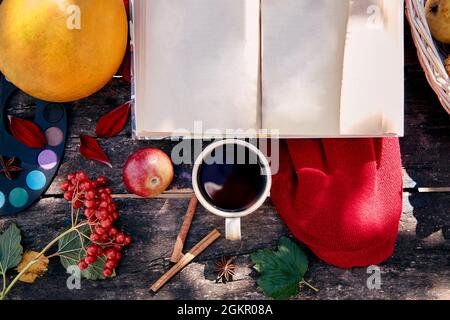 Cozy autumn concept with mug of tea, book mock up, paints and viburnum in rustic style. Autumn aesthetic still life with shadows, red dahlia. Cozy hom Stock Photo