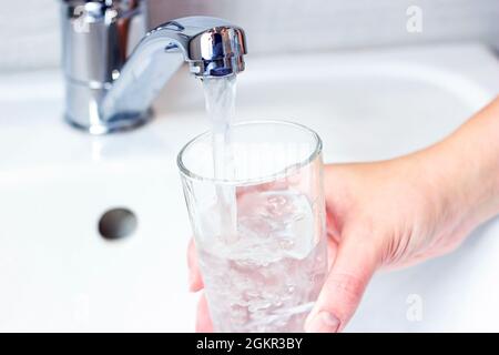 Young caucasian woman hand holding a glass with pure drinking water pouring from home faucet close up. Stock Photo