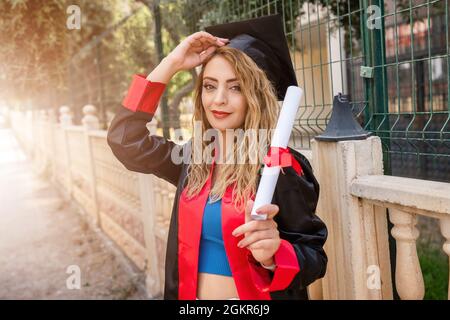 A pretty female student, celebrating her graduation and stand front of school fence, holding her diploma with success. High quality photo Stock Photo