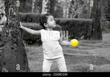 Grayscale shot of a little Southeast Asian girl playing with a yellow ball in a park Stock Photo