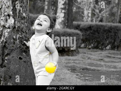 Grayscale shot of a little Southeast Asian girl playing with a yellow ball in a park Stock Photo