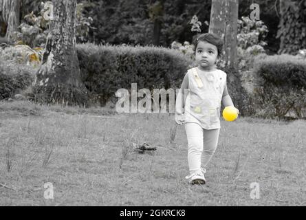 Grayscale shot of a little Southeast Asian girl playing with a yellow ball in a park Stock Photo