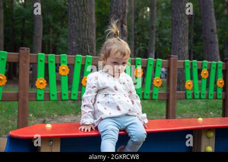 Cute adorable Caucasian little girl is playing on the outdoor playground. Girl in a white dress and hair braided in a ponytail and a sad face Stock Photo