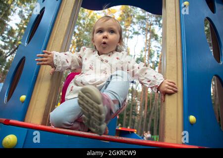 Cute adorable Caucasian little girl is playing on the outdoor playground. A girl in a white dress and her hair in a ponytail. Stock Photo