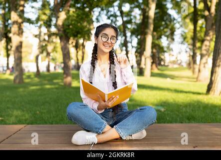 Happy indian female student writing in notebook outdoors, studying while sitting in campus park on bench Stock Photo