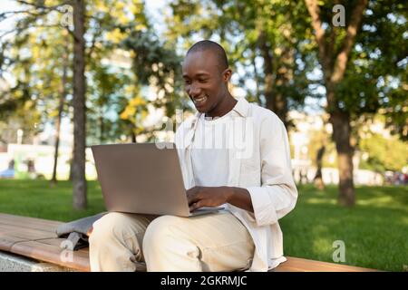 African american male student using laptop and learning online, doing homework outdoors in university campus park Stock Photo