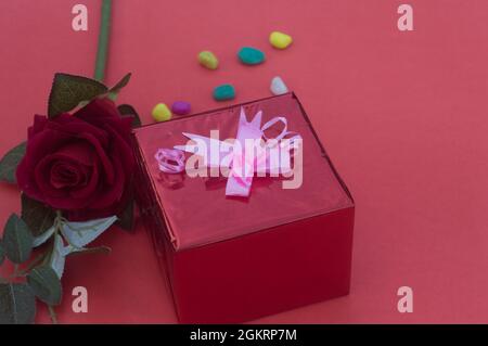 Close up gift box with tied bow ribbon placed near red rose on a table near in red background. High angle view. Design for valentine's day, anniversar Stock Photo
