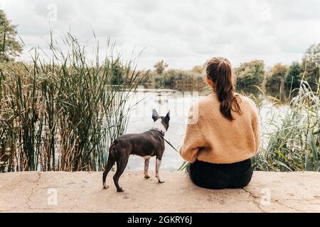 Teenage girl in a warm sweater  sitting with a Boston Terrier dog on the lakeshore - with her back turned, her dog staring at water - Autumn scenery Stock Photo
