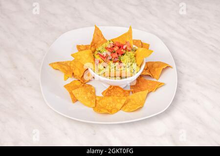 Small white bowl of guacamole and pico de gallo with cornmeal nachos for dipping in white plate on white marble table Stock Photo