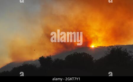 Wildfire on a mountain slope at sunset with dramatic red sky, heavy smoke and silhouette of helicopter with water bucket Stock Photo