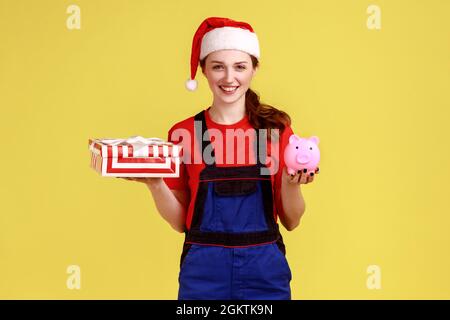 Happy delivery woman holding wrapped gift box and piggy bank, savings for online delivering service, wearing blue overalls and santa claus hat. Indoor Stock Photo