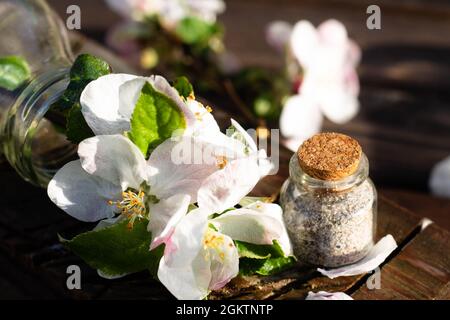 Transparent glass bottle with water and apple tree flowers on a rustic wooden tabletop with a small jar of sea sand. Dark background Stock Photo