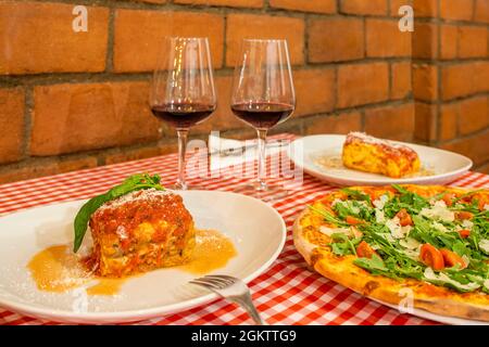 Table in the dining room of an Italian restaurant with pizza to share and individual plates of lasagna and two glasses of red wine to soak your throat Stock Photo