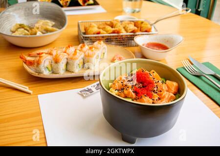 Black bowl with marinated salmon, fish roe, avocado chunks and uramaki rolls with prawns and gyozas on the bottom Stock Photo