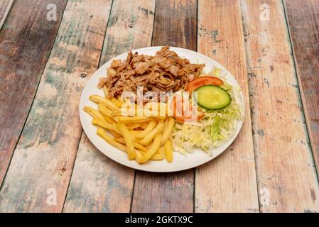 Kebab plate garnished with fried potatoes with roast lamb meat with iceberg lettuce salad, tomato slices and cucumber on wooden table Stock Photo