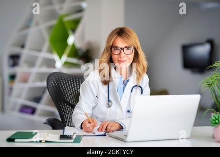 Smiling female healthcare worker text messaging and using laptop while working at doctor's office. Stock Photo