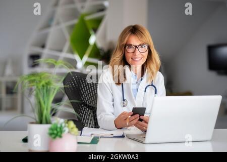 Smiling female healthcare worker doing some paperwork and using laptop while working at doctor's office. Stock Photo
