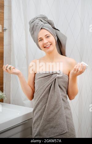 Woman in towel after a shower in the bathroom holds a white jar of cream. Stock Photo