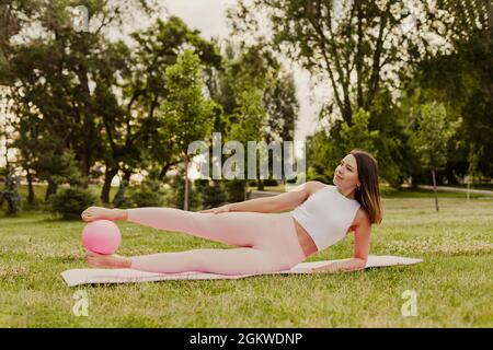 attractive, elegant woman is engaged in pilates with ball on green grass of lawn at sunset Stock Photo