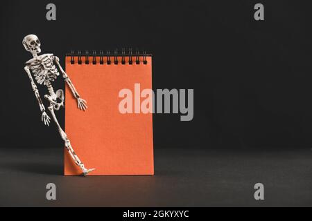 Blank orange notebook on black background, with skeleton. Banner for congratulations or invitations. Minimalistic holiday design for Halloween party. Stock Photo