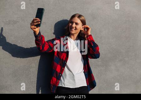 Excited young woman, taking a selfie on a mobile phone, wearing casual fashionable clothes, walking in the city against the background of a gray biton Stock Photo