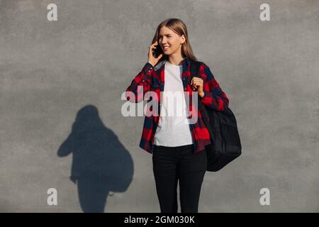 Portrait of a smiling stylish young woman, in a red plaid shirt, with a backpack on his shoulder, a university student talking on a mobile phone, a fe Stock Photo