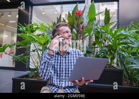 Millennial businessman talks on the phone while holding up his laptop. Smiling successful person at work - elegant caucasian man during a phone call. Stock Photo