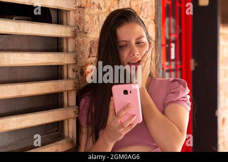 Surprised woman looks at her pastel pink cell phone, wearing pastel pink blouse Stock Photo