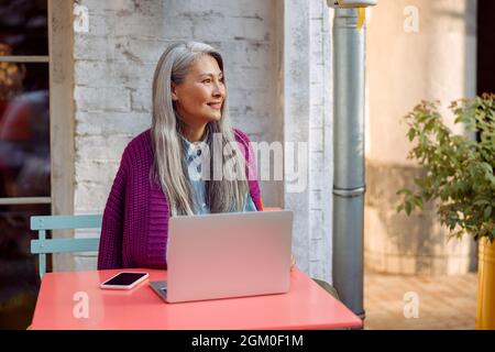 Senior Asian lady in knitted jacket with smartphone and laptop sits at table on terrace Stock Photo