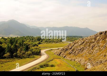 A gravel road goes through the valley, bending around the slope of the gova along the edge of a dense forest. Karakol Valley, Altai, Siberia, Russia. Stock Photo