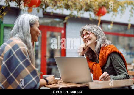 Grey haired lady with laptop spends time with Asian friend on outdoors cafe terrace Stock Photo