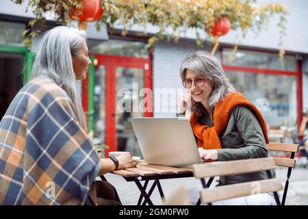 Hoary haired Asian woman and mature friend with laptop on outdoors cafe terrace Stock Photo