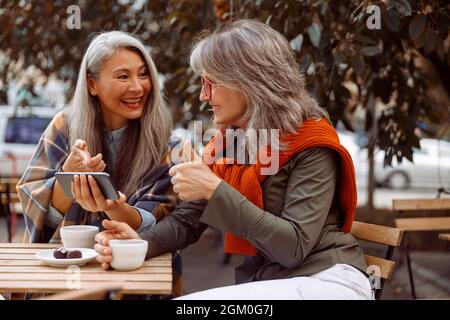 Cheerful senior Asian lady shows phone to grey haired friend at table with coffee and candies Stock Photo