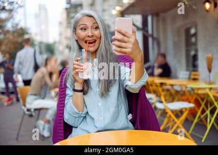 Joyful hoary haired Asian lady takes selfie with smartphone on outdoors cafe terrace Stock Photo