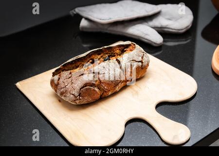 Rustic baked sourdough bread on cuttings board placed on black kitchen counter. Oval fresh yeast bread loaf made with rye and wheat flour and nuts Fro Stock Photo