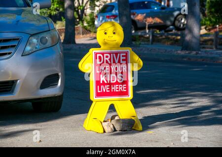 Drive Like Your Kids Live Here road sign in residential neighborhood near parked car. Child like yellow figure with red safety sign on road. Stock Photo
