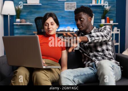 Married interracial couple looking at modern laptop on couch. Multi ethnic partners using digital device with technology, watching display of gadget. Mixed race people with computer Stock Photo
