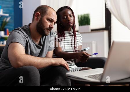 Interracial couple using laptop for taxes payment and economy at home. Mixed race people doing accounting for financial budget and tax money while using device with technology. Stock Photo
