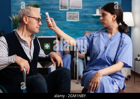 Support assistant worker measuring disabled senior man temperature using medical infrared thermometer during medicine therapy. Social services nursing elderly retired male. Healthcare assistance Stock Photo