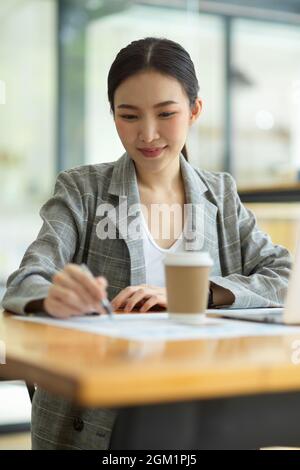 Beautiful young businesswoman using laptop and doing some paperwork, writing down her ideas, drinking coffee Stock Photo