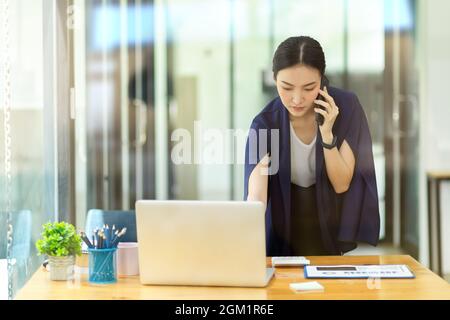 Businesswomen standing in office, talking on smart mobile phone and typing on laptop keyboard. multitasking businesswomen, hard working women, female Stock Photo
