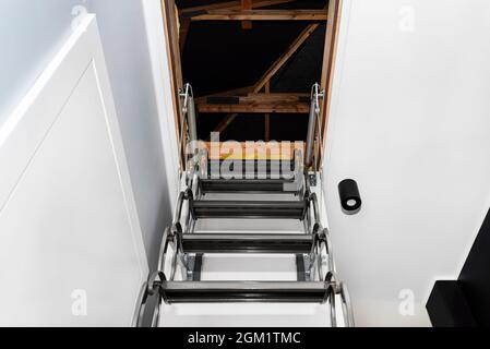 Metal stairs hidden in the ceiling to the attic with an opening hatch and folding stairs in the corridor, view from the bottom of the stairs up, moder Stock Photo