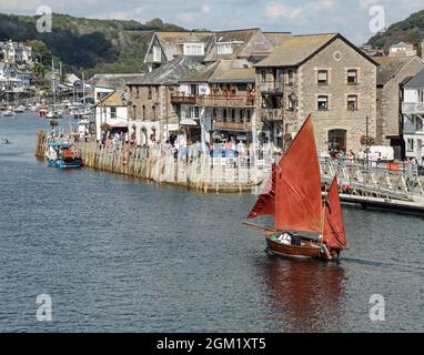 Iconic red canvas sails on a boat travelling up the River Looe in Cornwall with a busy quayside at East Looe as a backdrop. Looe Cornwall. Stock Photo