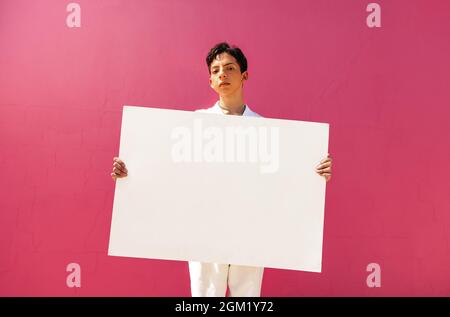 Confident queer boy holding a blank placard against a pink background. Assertive teenage activist displaying a white banner in a studio. Young gay boy Stock Photo