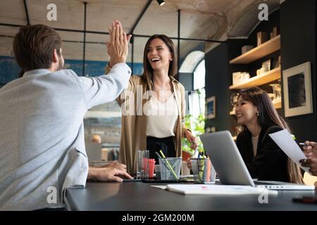 Two cheerful young business people giving high-five while their colleagues looking at them and smiling. Stock Photo