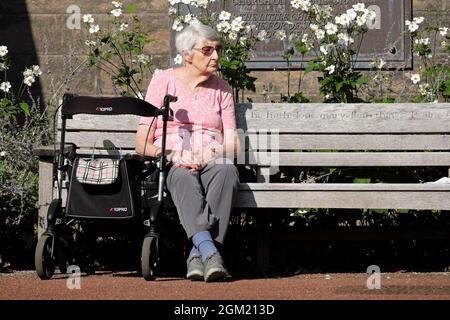Elderly woman sat on church bench in the sunshine with a walking frame aid September 2021 UK Stock Photo