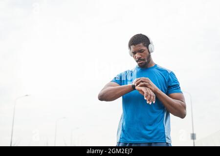 Young muscular man in wireless headphones using fitness bracelet during sports training outdoors Stock Photo