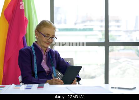 A senior designer uses her tablet computer to research information for modern design. A clothes rack with colorful fabrics in front of the large windo Stock Photo